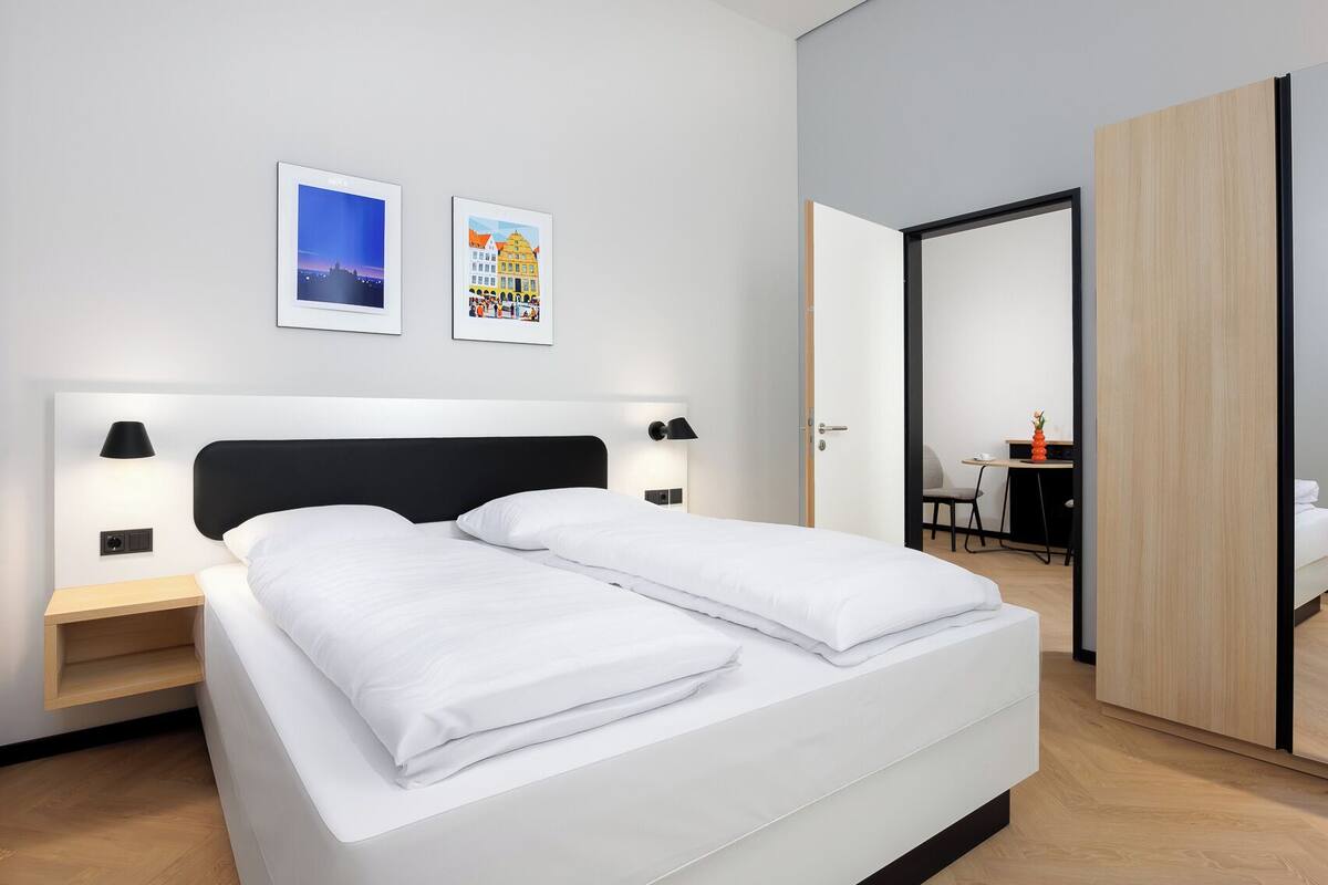 Una habitación de hotel moderna con una cama grande, dos mesitas de noche con lámparas, un armario de madera y un escritorio con una silla.