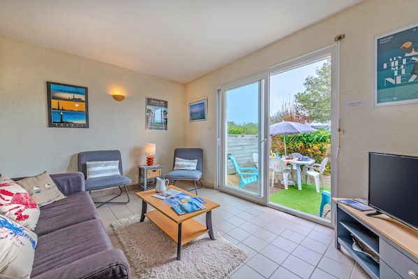 A living room with a sofa, two armchairs, a coffee table, a TV, and a sliding glass door leading to an outdoor area with a table and chairs.