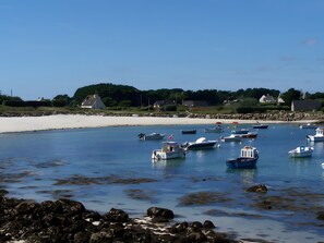 Un paysage côtier avec des bateaux ancrés dans des eaux calmes, une plage de sable et des maisons en arrière-plan.