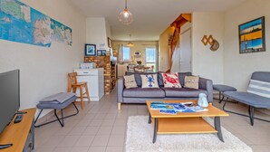 Een moderne woonkamer met een bank, salontafel en televisie. Achtergrond toont een keuken met een eethoek.