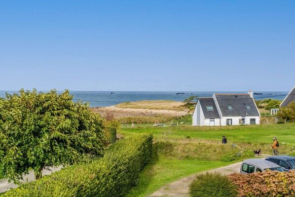 A coastal property with a pathway leading to a white house, a parked car, and a dog walking.