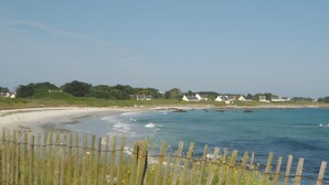 Een kustgezicht met een zandstrand, een houten omheining en huizen op de achtergrond.