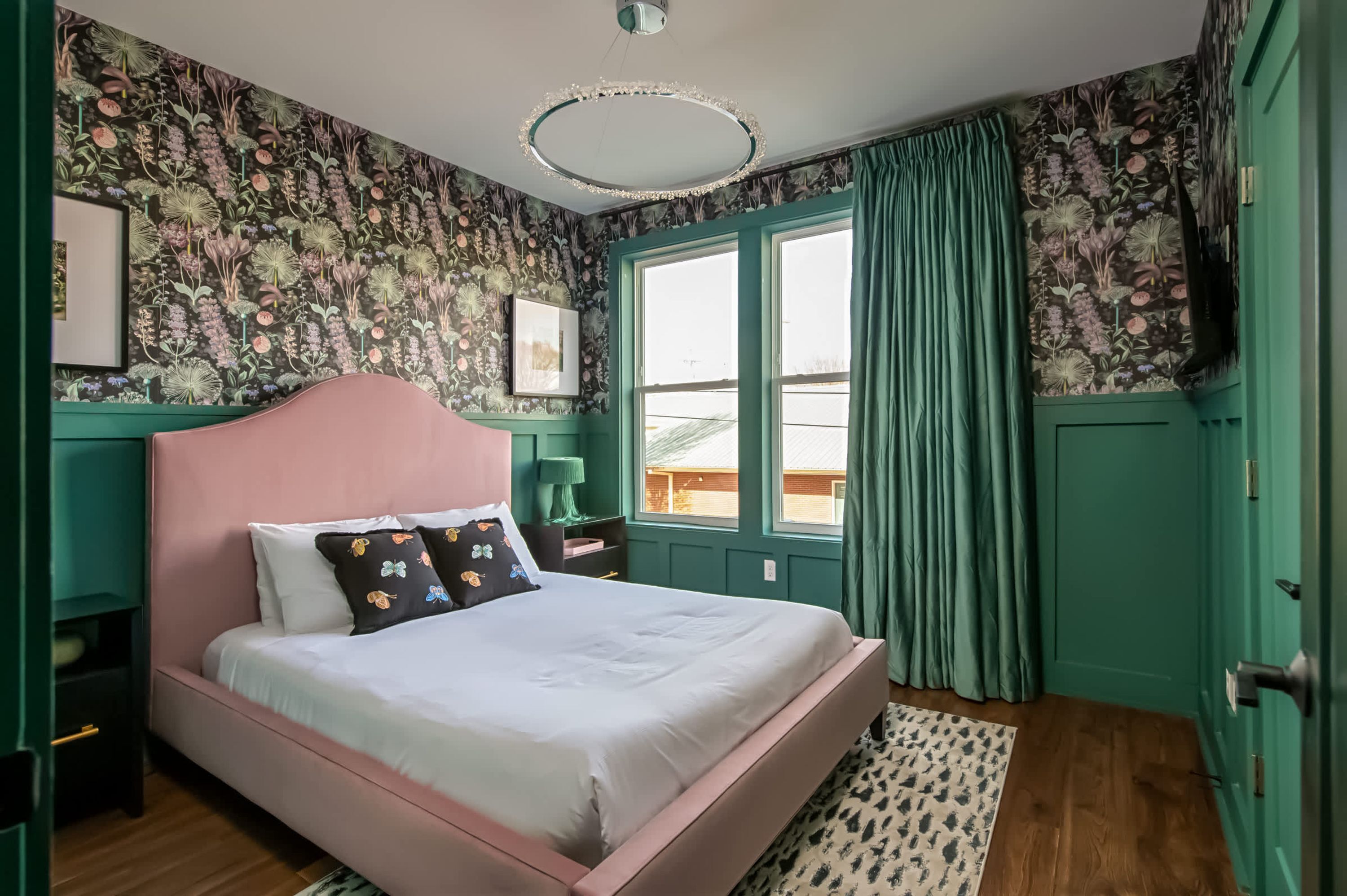 Une chambre avec un lit rose, un papier peint à motifs floraux, une porte verte et une fenêtre avec des rideaux.