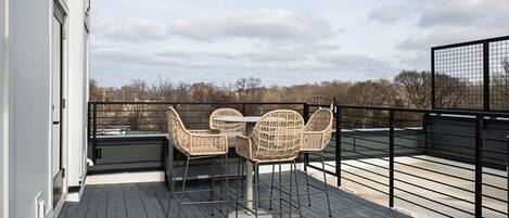 A rooftop terrace with wicker chairs and a table, overlooking a cityscape.