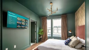 A modern bedroom with a large bed, a TV mounted on the wall displaying a menu, and a window with curtains. - Rooftop Hot Tub Skyline Views Near Broadway (Nashville)