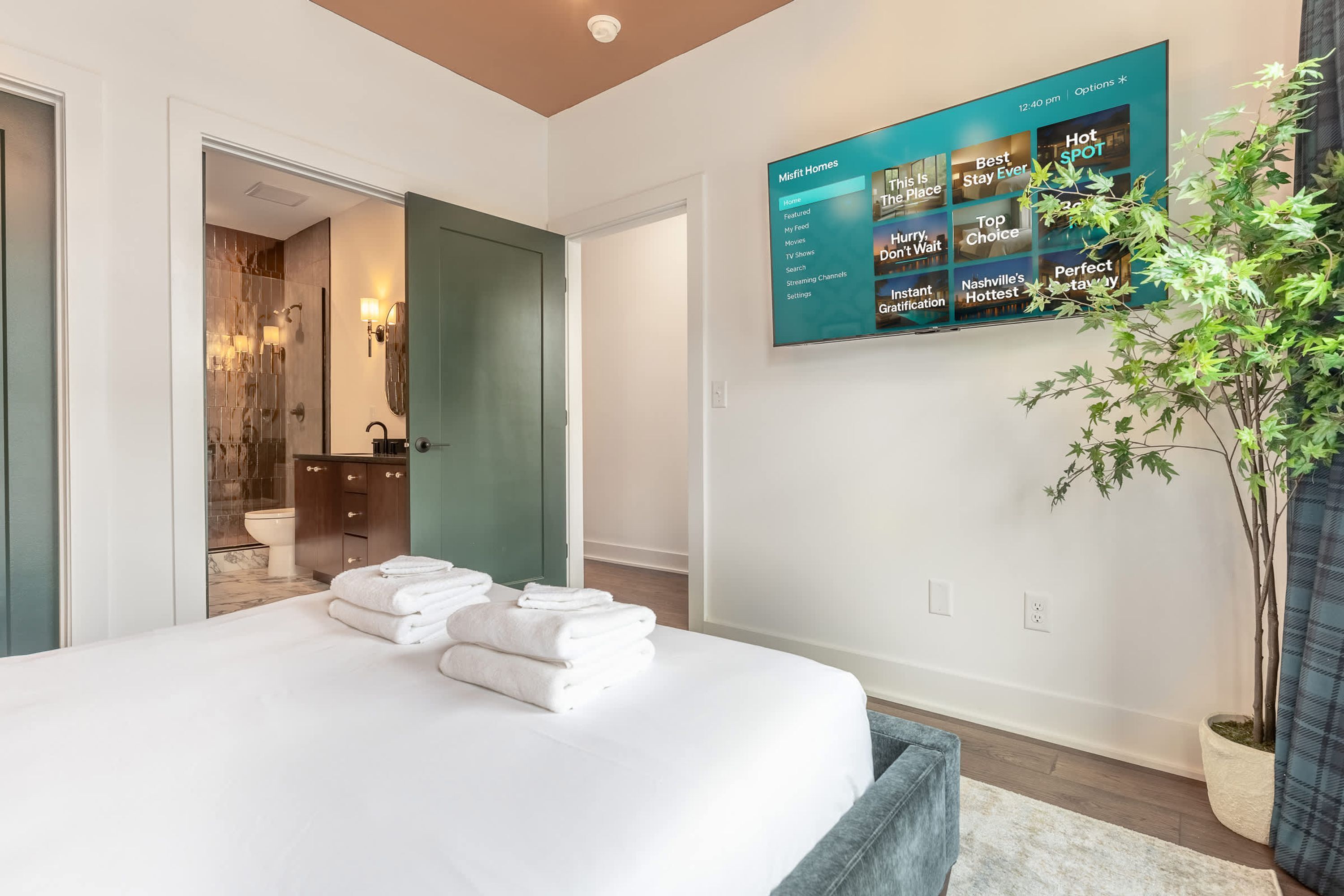 Une chambre d’hôtel moderne dotée d’un grand lit, d’une salle de bain avec une douche en verre et d’un téléviseur fixé au mur qui diffuse différentes chaînes.