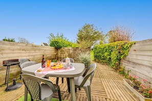 Une terrasse en bois avec une table dressée pour deux personnes, entourée de chaises, et un barbecue situé à gauche.