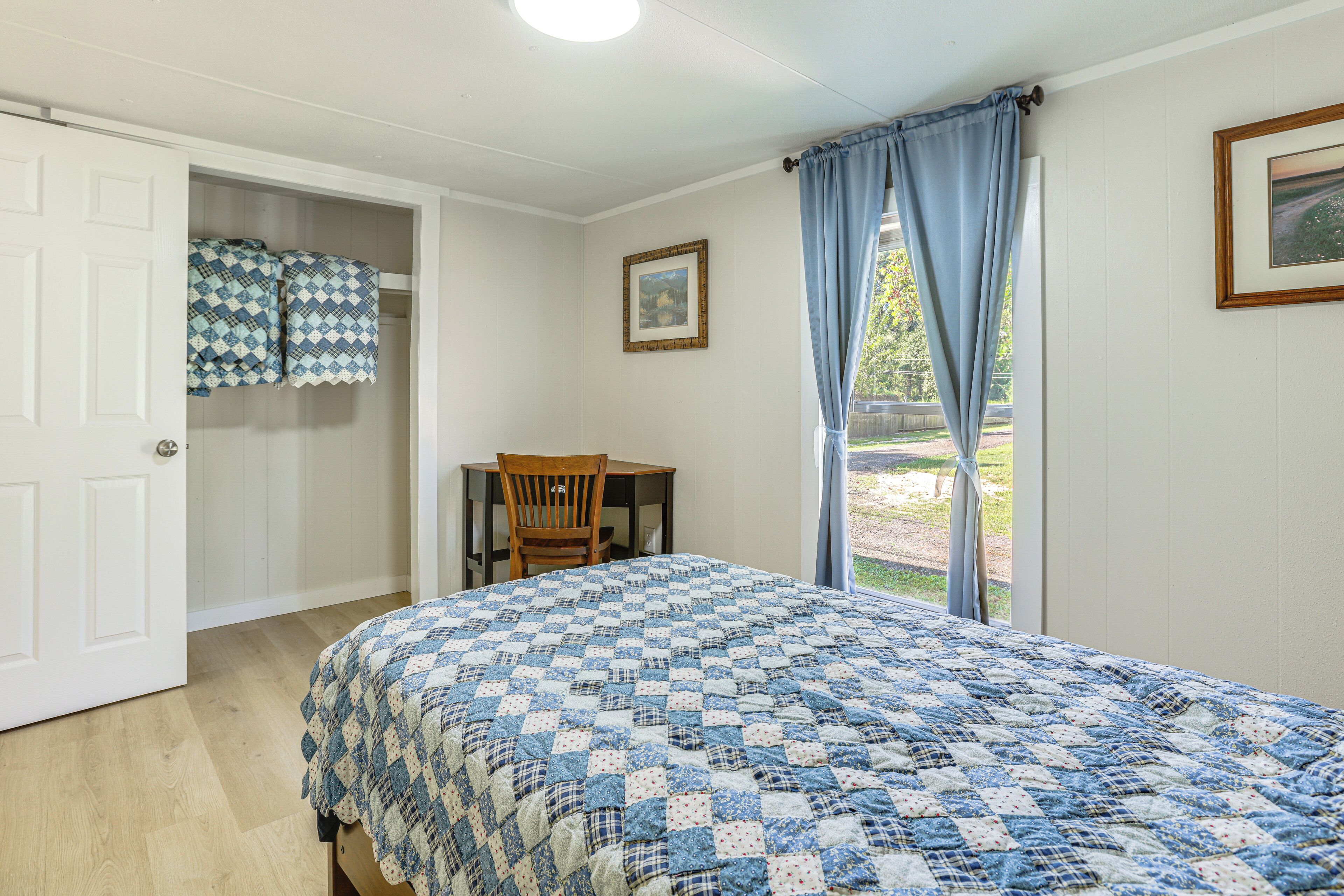 Un dormitorio con una colcha acolchada, una silla de madera, un escritorio y una ventana con cortinas.