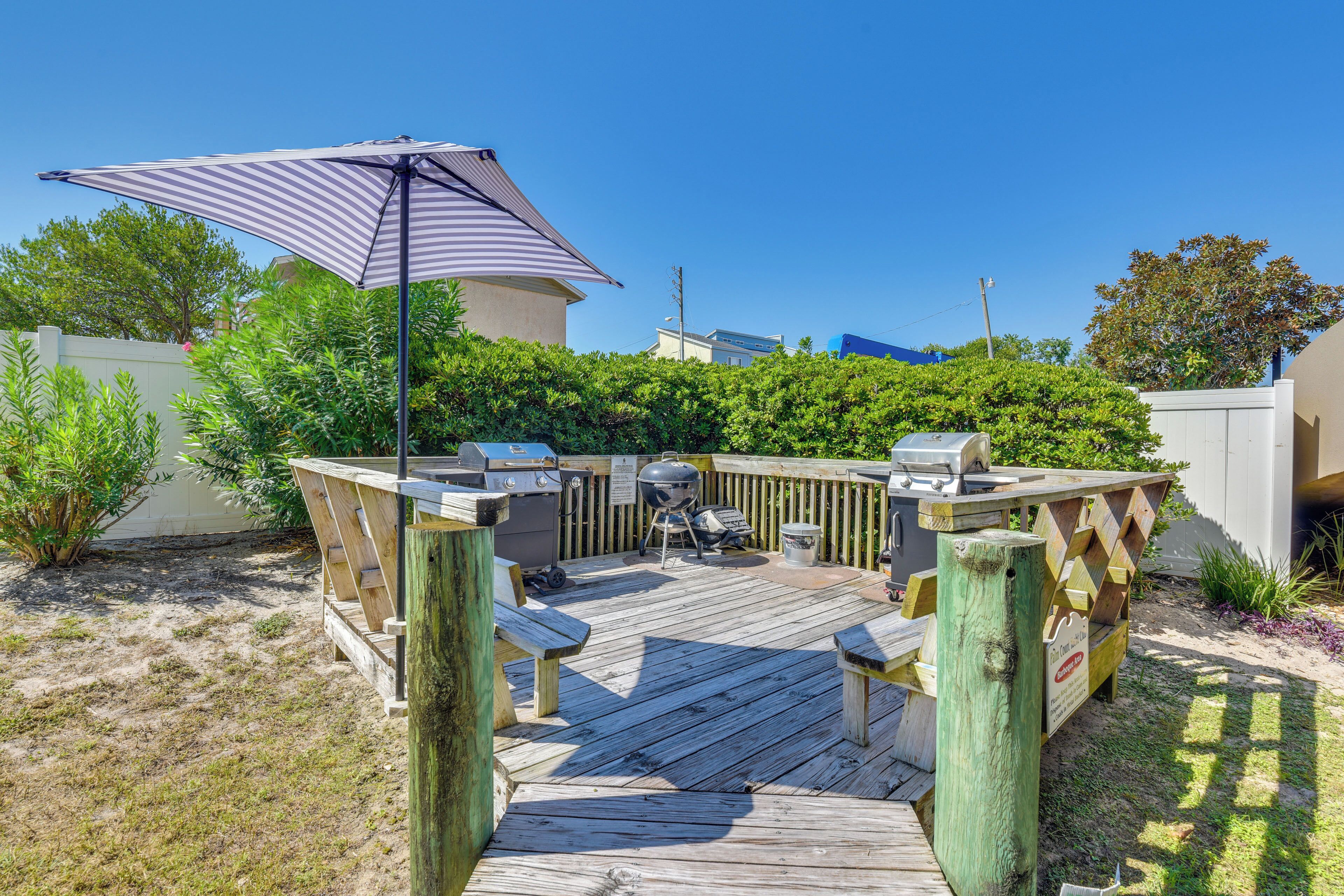 Une terrasse en bois avec un barbecue et un espace salon, entourée de verdure et sous un ciel bleu dégagé.