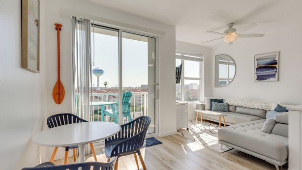 A modern living room with a sofa, a dining table with chairs, a ceiling fan, and a view of buildings through large windows. - Walk to Sand! Modern Okaloosa Island Condo (Fort Walton Beach)