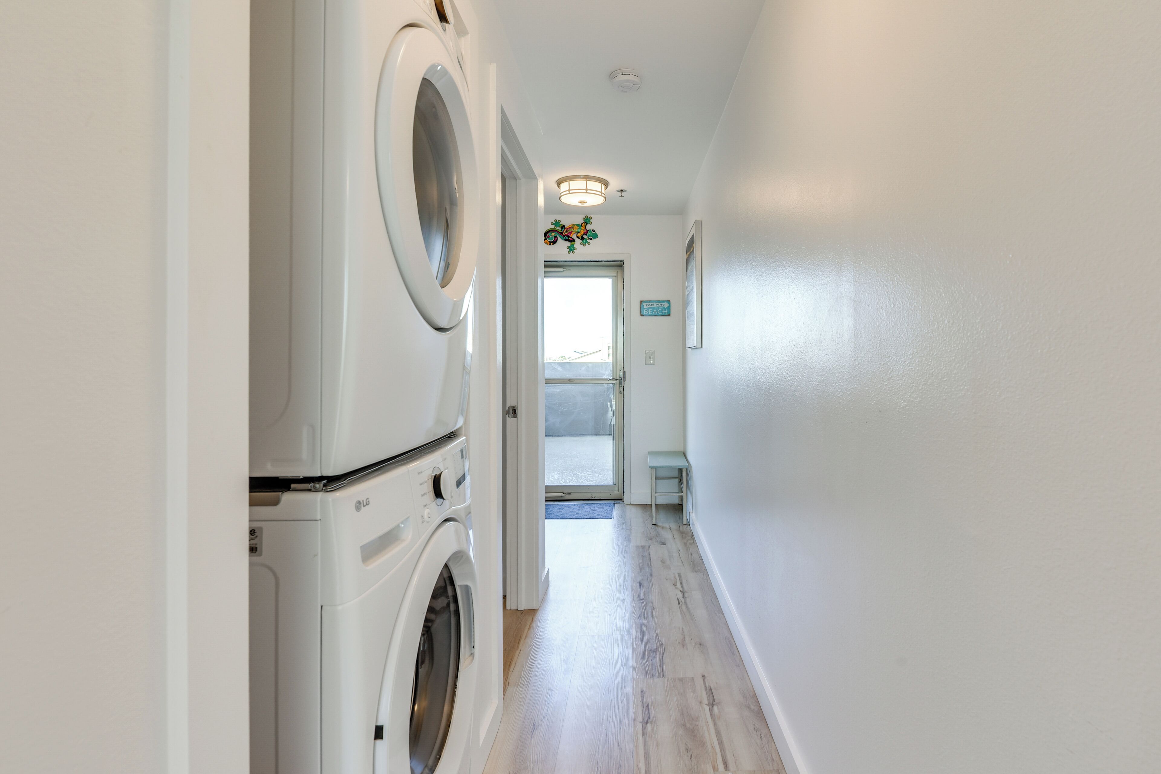 A laundry room with a front-loading washer and dryer, a hallway leading to a door with a view of the outdoors, and a small table with a sign that reads 'THIS WAY BEACH'.
