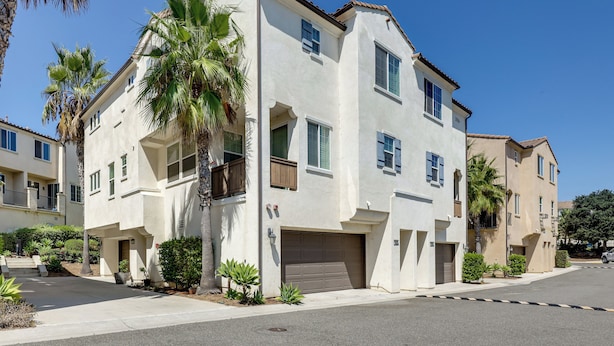 A two-story residential building with a garage, surrounded by palm trees and other greenery. - Walk to Public Transit: Gated Oceanside Abode! (Oceanside)