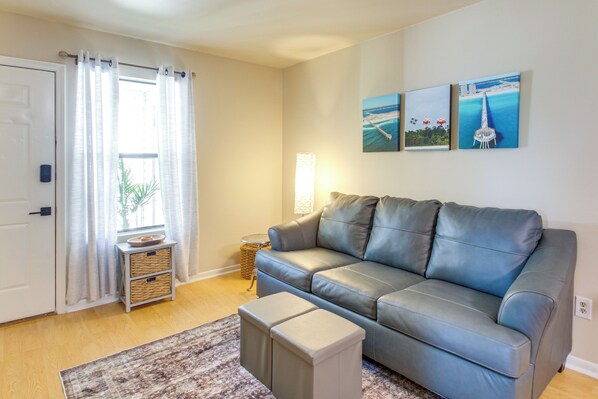 Ein Wohnzimmer mit grauem Ledersofa, einem kleinen Ottomanen, einem gemusterten Teppich, einer Tür mit weißem Vorhang und drei gerahmten Bildern an der Wand.