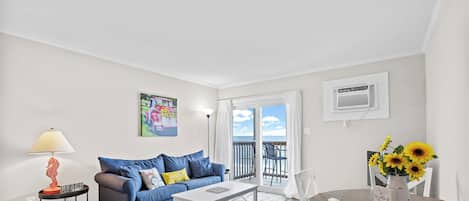 Una sala de estar con un sofá azul, una mesa de centro blanca y una mesa de comedor con sillas blancas. Hay un cuadro en la pared, una lámpara sobre una mesita auxiliar y vistas al exterior a través de una puerta corrediza.