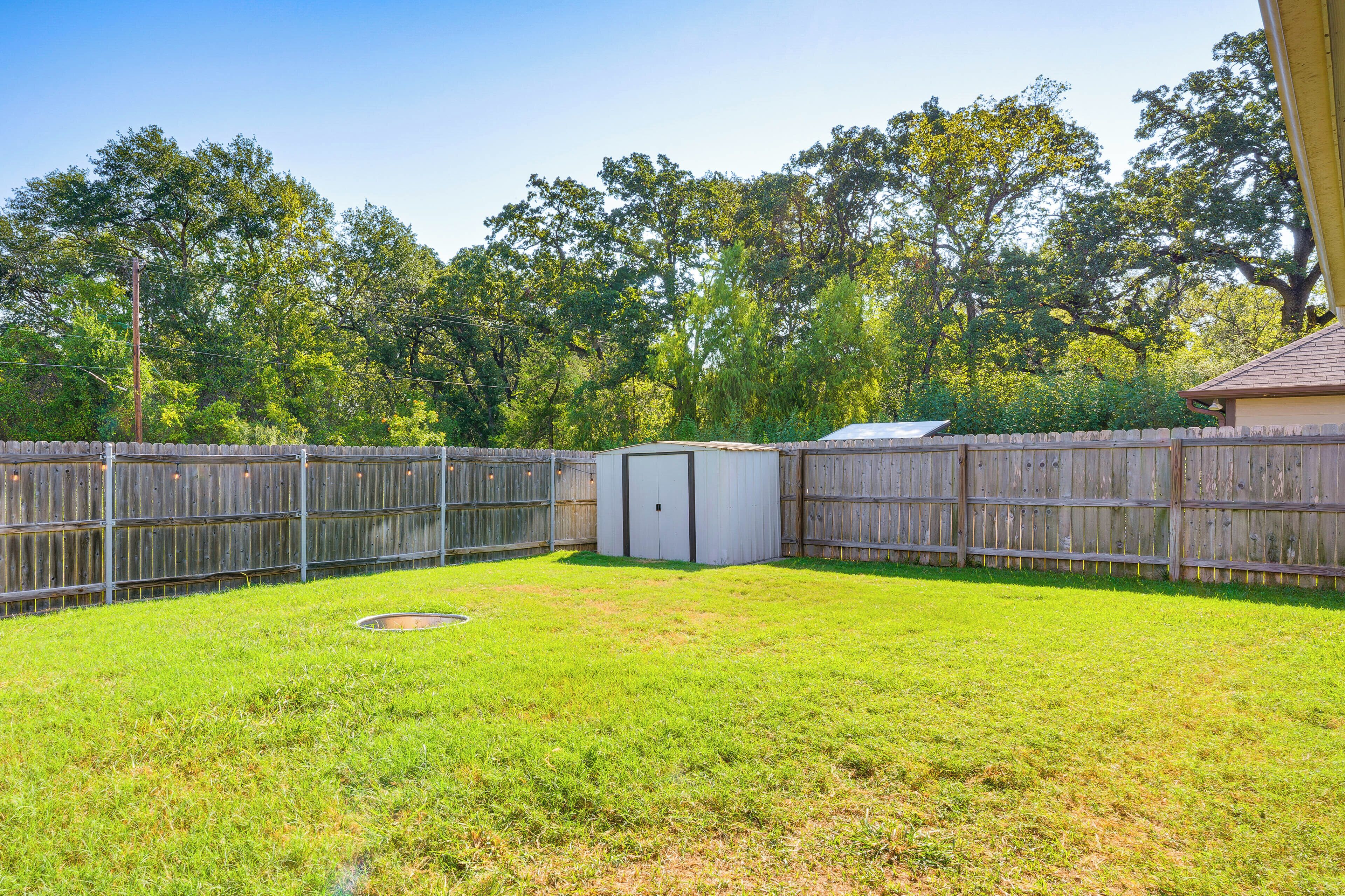 A fenced backyard with a small shed, a grassy lawn, and trees in the background.