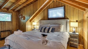 A cozy wooden cabin bedroom with a large bed, a window, and a small table with a lamp. - < 1 Mi to Lake: Cabin w/ Hot Tub in Greentown! (Greentown)
