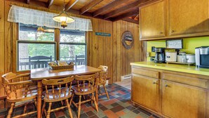 A wooden cabin interior with a dining table and chairs, a kitchen area with a yellow countertop, and a window with a view of trees. - < 1 Mi to Lake: Cabin w/ Hot Tub in Greentown! (Greentown)