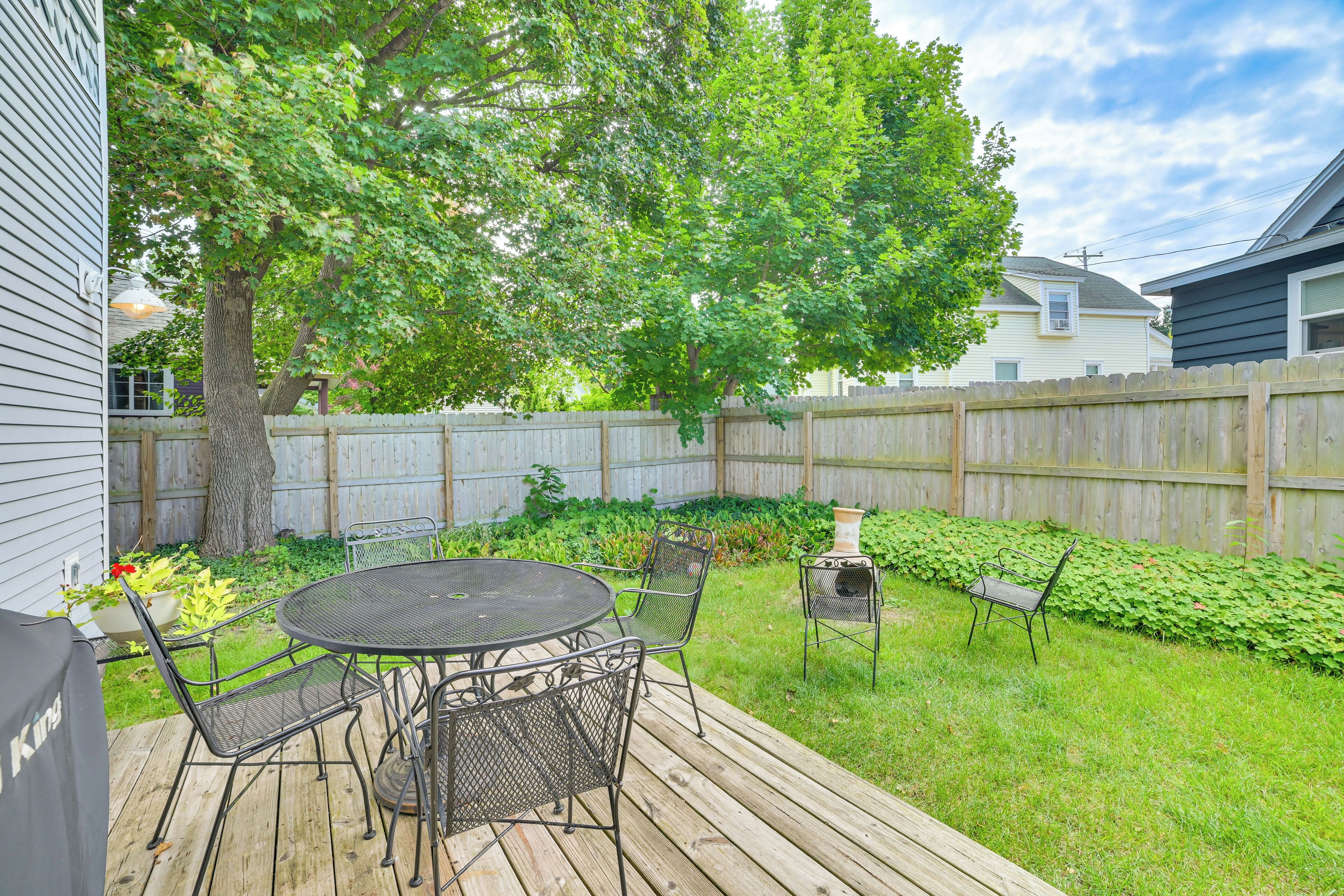 Un jardin avec une terrasse en bois, une table ronde entourée de chaises, un barbecue et une clôture en bois.