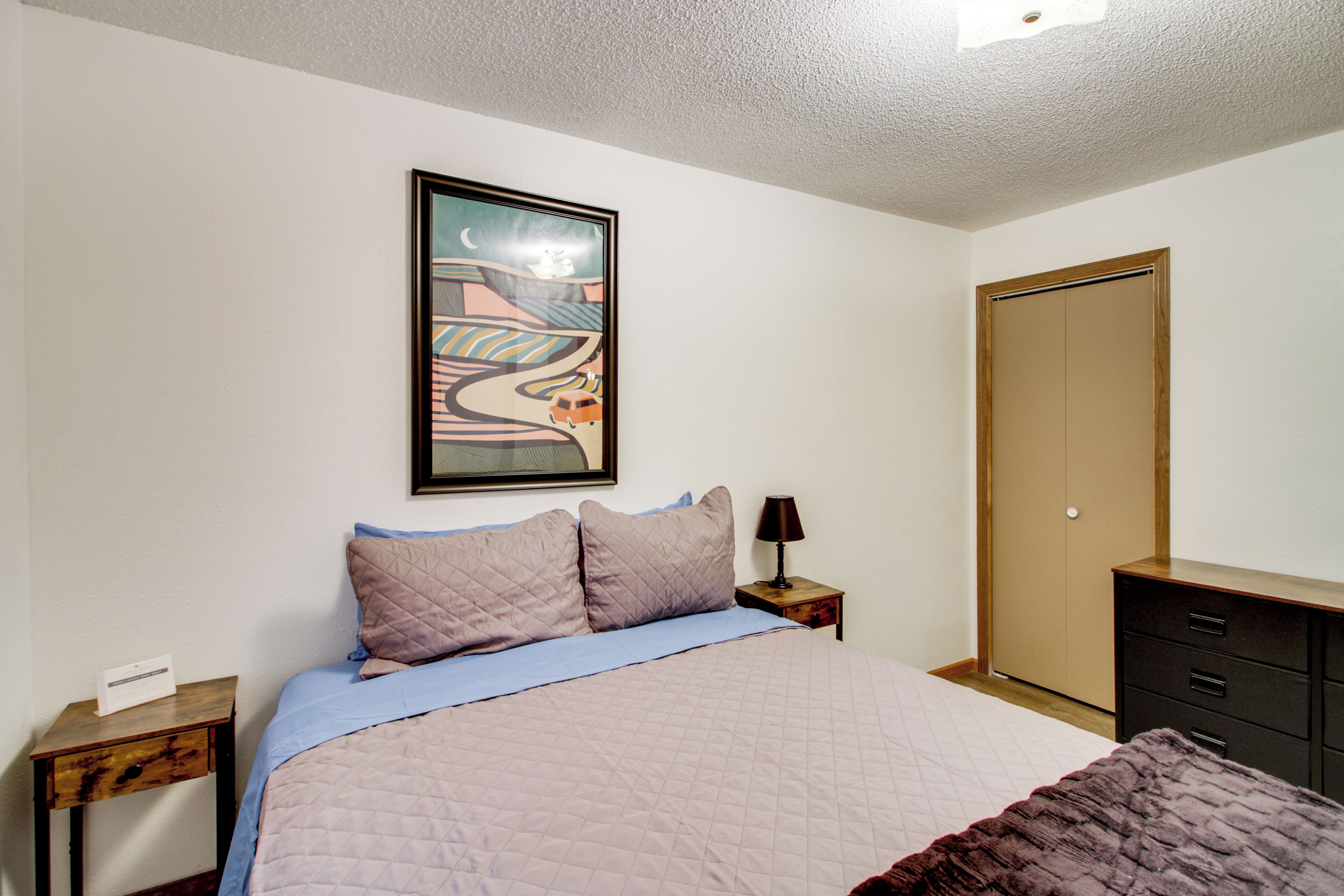A bedroom with a bed, a nightstand, a dresser, a wardrobe, and a framed picture on the wall.