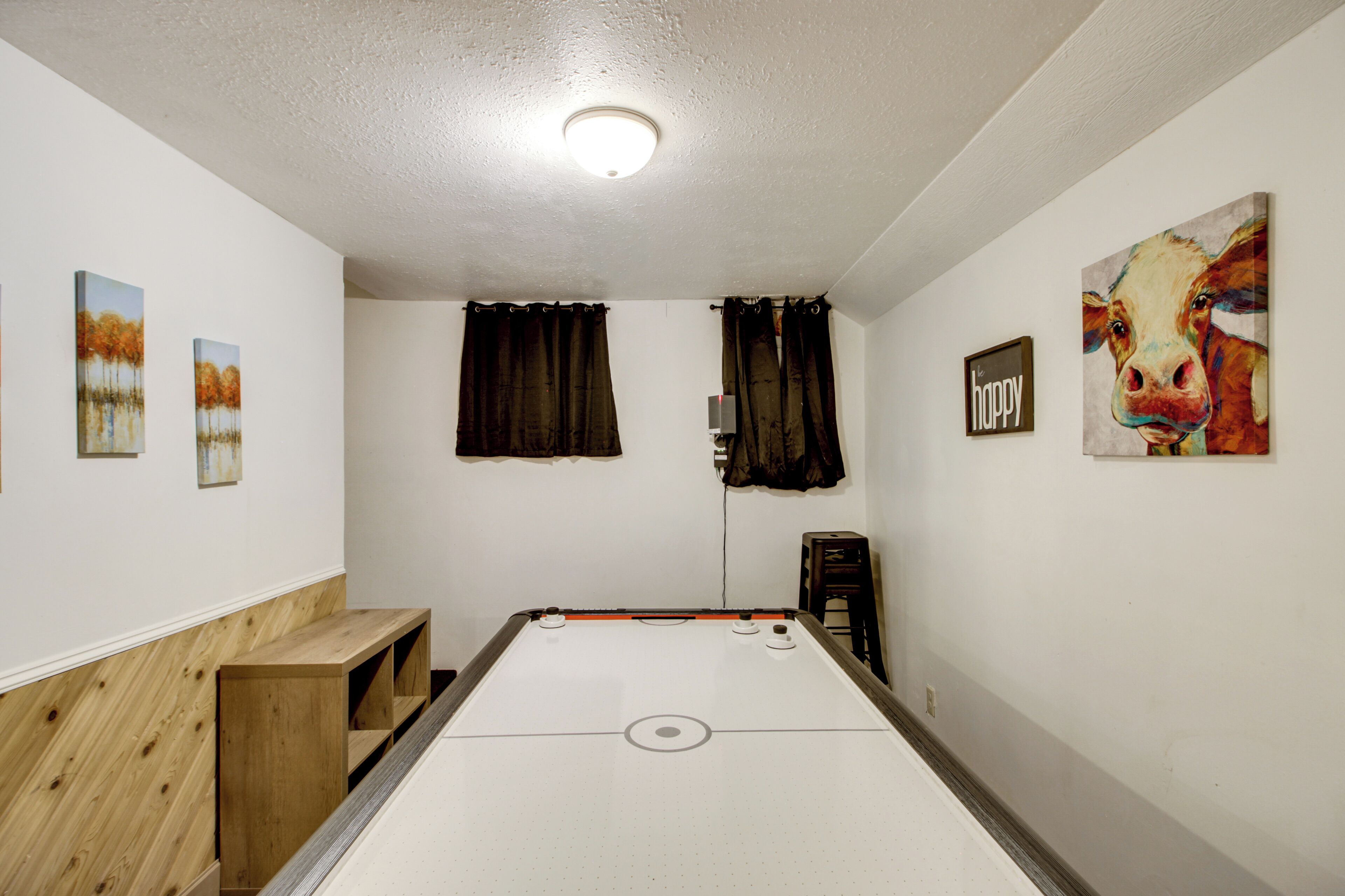 A pool table in a room with white walls, a ceiling light, and artwork including a painting of a cow and three landscape paintings.