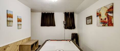 A pool table in a room with white walls, a ceiling light, and artwork including a painting of a cow and three landscape paintings.