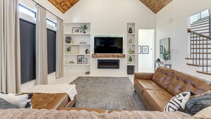 A modern living room with a fireplace, a large TV, and a staircase. - Luxe Retreat w/ Patio & Fenced Yard in Waxahachie! (Waxahachie)