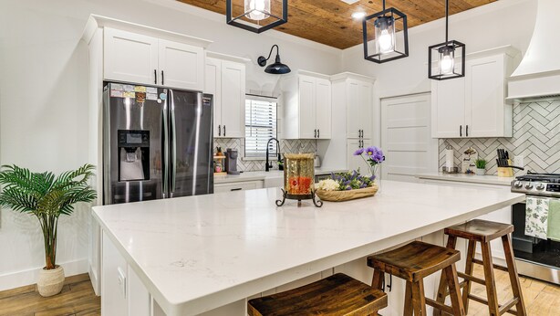 Una cocina moderna con armarios blancos, electrodomésticos de acero inoxidable, encimera de mármol, taburetes de madera e iluminación colgante.