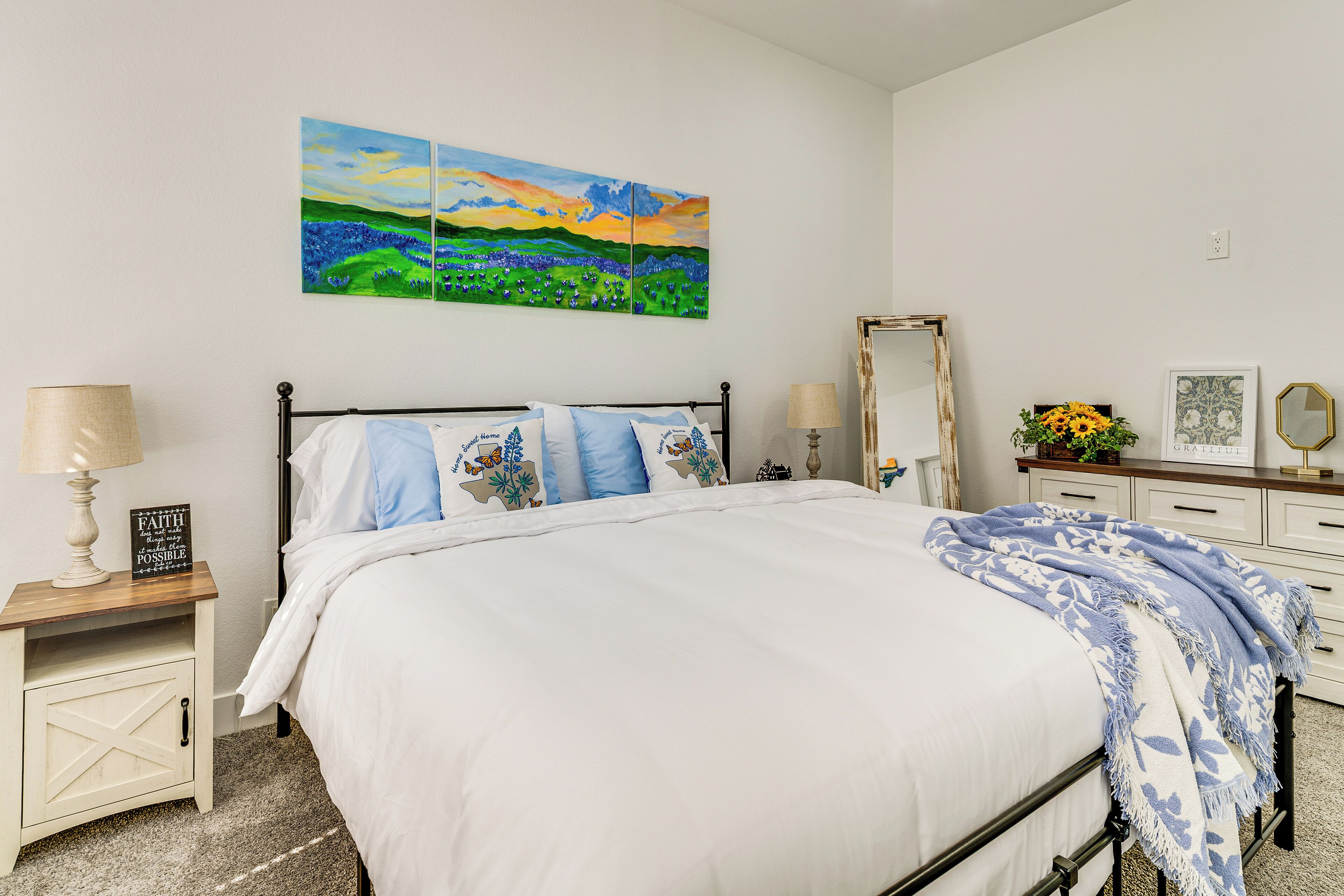 Een netjes opgemaakt bed met een witte dekbedovertrek en blauw-witte kussens, een nachtkastje met een lamp, een lijst met spiegel en een klein tafeltje met een vaas zonnebloemen.