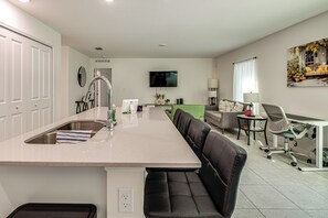 Een moderne keuken met een centraal eiland, een zithoek met een bank en stoelen, een aan de muur bevestigde televisie en een raam met gordijnen.