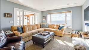 A spacious living room with a large sectional sofa, a leather ottoman, and a view of the ocean through a large window. - Pool & Stunning Water Views: Kent Island Home! (Stevensville)