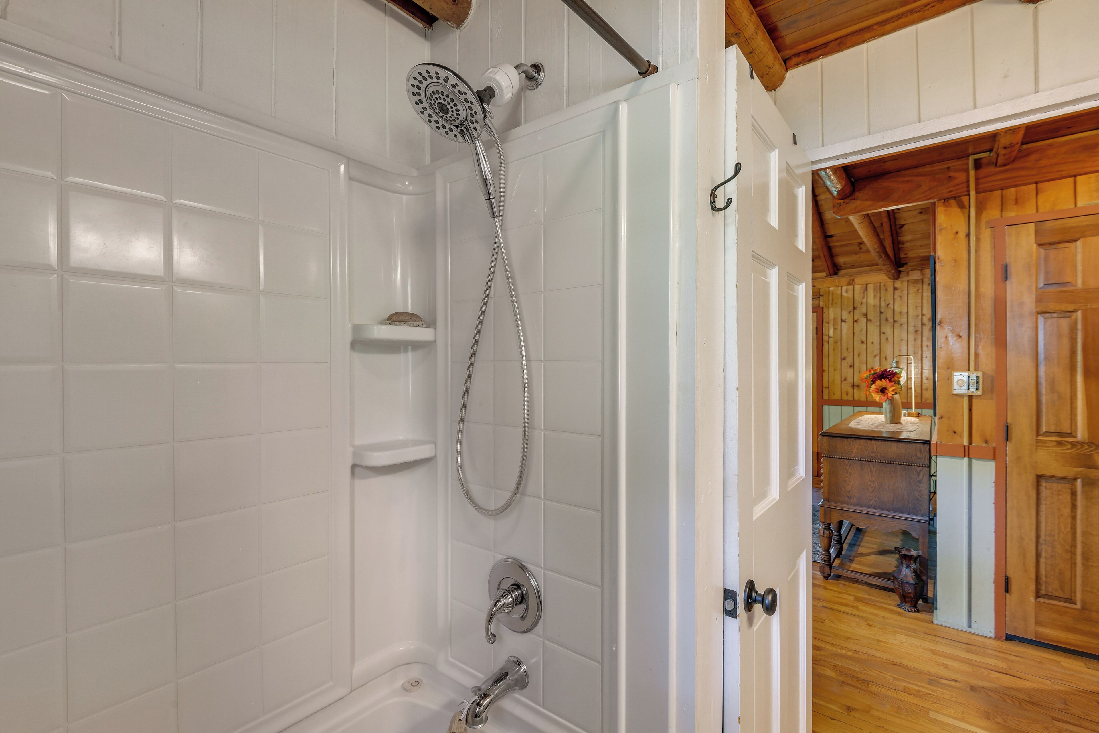 A bathroom with white tiled walls, a shower with a handheld showerhead, and a wooden door leading to a hallway with a wooden floor.