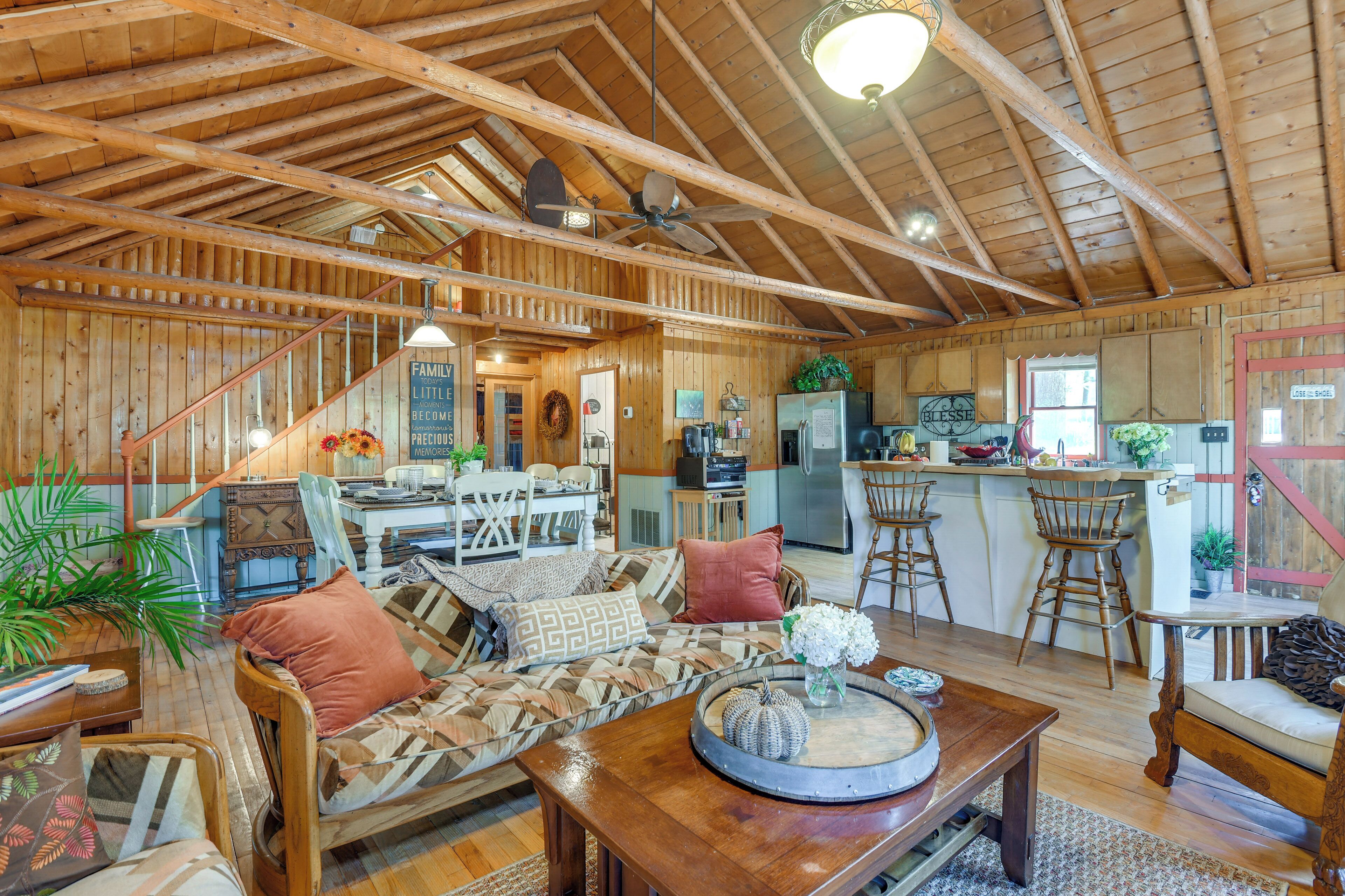 A spacious living area with a high ceiling, wooden beams, a dining table, and a kitchen area with a refrigerator and bar stools.