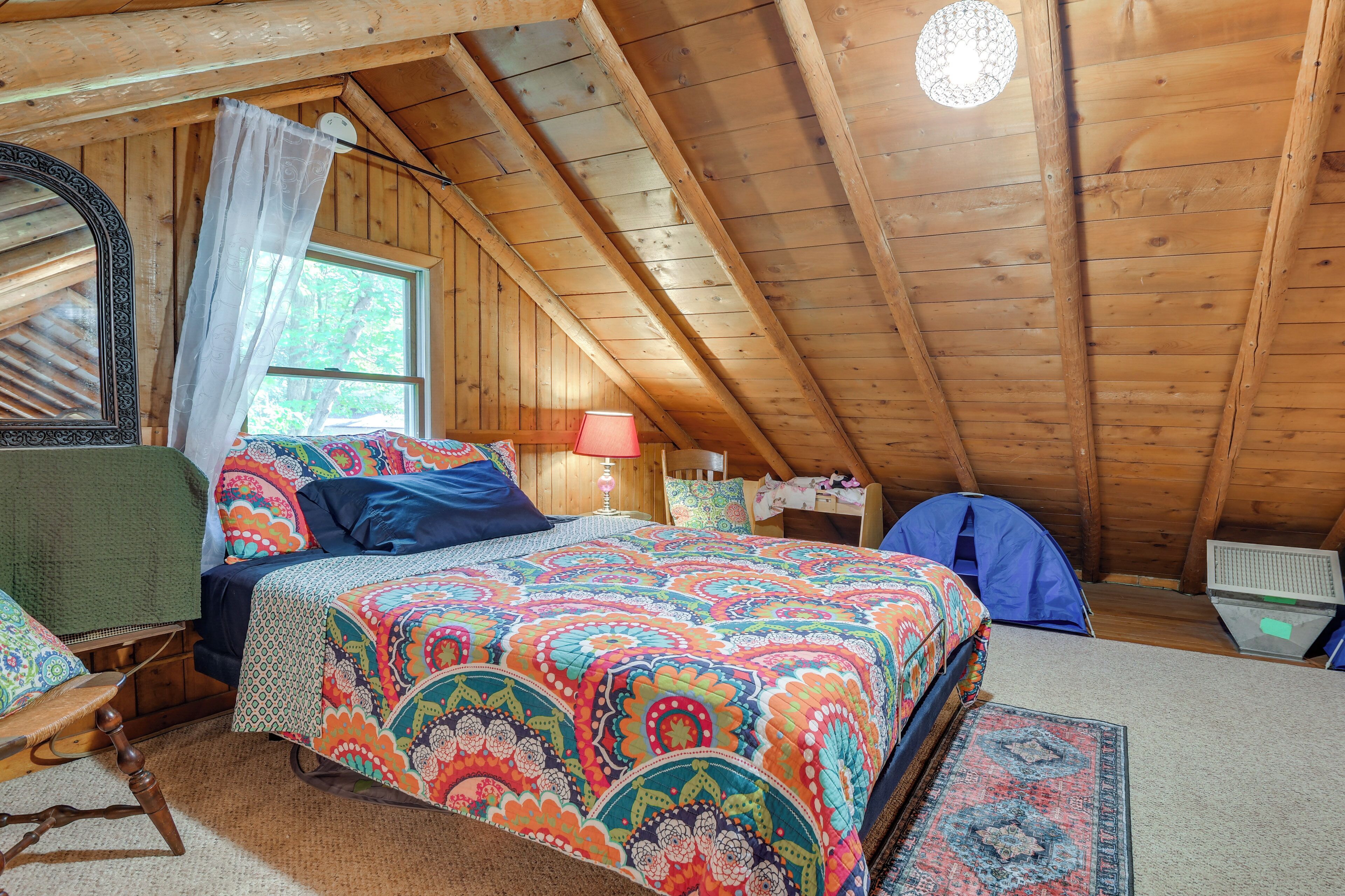 A cozy attic bedroom with a colorful bedspread, a wooden ceiling, and a small desk with a lamp.