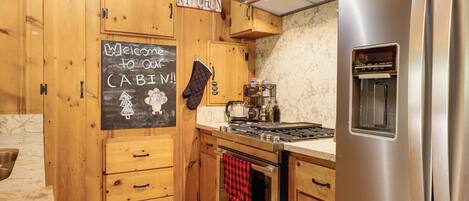 A kitchen with wooden cabinets, a stainless steel refrigerator, a stove, and a chalkboard with a welcome message.