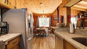 A wooden cabin kitchen with a refrigerator, coffee maker, wooden dining table, chairs, and a window with a view of trees. - Community Lake Access: Peaceful Cabin in Sonora! (Sonora)