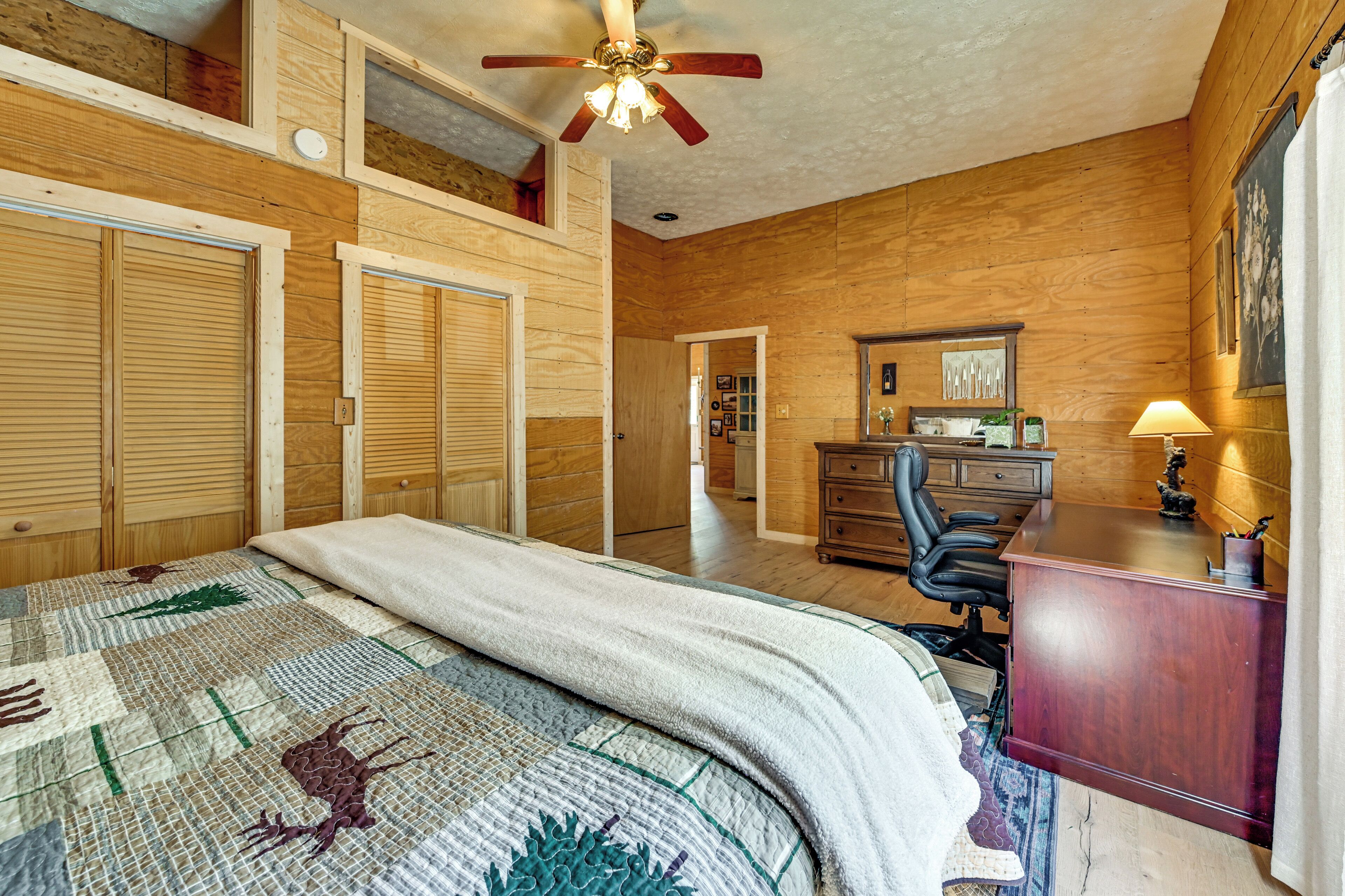 A bedroom with wooden walls, a bed with a quilt, a desk with a lamp, and a ceiling fan.