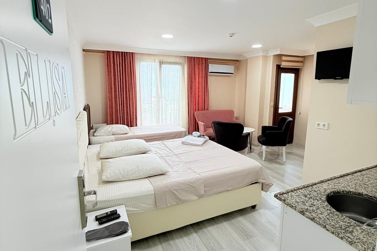 A hotel room with a large bed, a desk, a chair, a TV, and a kitchenette.
