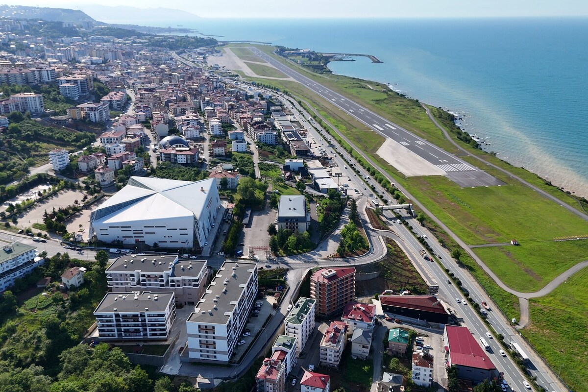 Vista aérea de una ciudad costera con un aeropuerto destacado, edificios residenciales y un gran edificio blanco con una estructura de techo singular.