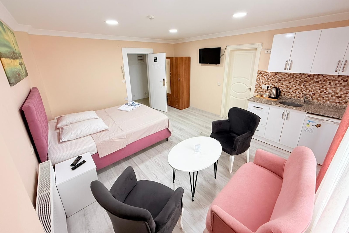 A compact hotel room with a bed, a desk, a chair, a small table, a kitchenette, and a TV.