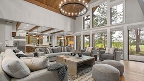 A spacious living room with a large gray sectional sofa, a coffee table, and gray poufs. There is a kitchen area with a refrigerator and a dining area with a table and chairs. The room has large windows offering a view of the outdoors. - Luxury Family Escape with Access to Forest House (Sunriver)