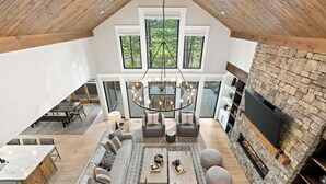 A spacious living area with a stone fireplace, a large chandelier, and a view of the outdoors through large windows. - Luxury Family Escape with Access to Forest House (Sunriver)