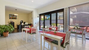 Un espace repas avec des tables et des chaises blanches, une plante en pot et une cuisine en arrière-plan.