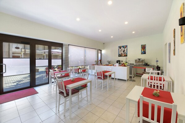 Un espace repas avec des tables blanches et des chaises rouges, un coin cuisine et une vue dégagée grâce à de grandes portes vitrées.