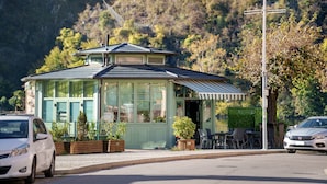 Un edificio pequeño con techo verde, amplias ventanas, un porche y un comedor con mesas al aire libre.