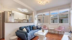 A modern living room with a sofa, coffee table, and chairs, a kitchen area with cabinets, and large windows. - La Justicia Apartment with Private Parking, Shared Terrace, Wi-Fi & Air Conditioning (Jerez de la Frontera)