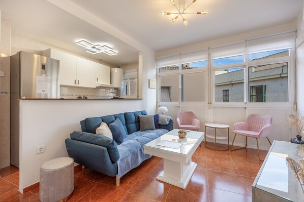 A modern living room with a sofa, coffee table, and chairs, a kitchen area with cabinets, and large windows.