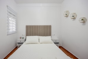 A bedroom with a bed, two bedside tables, a window with blinds, and three wall-mounted decorative items.