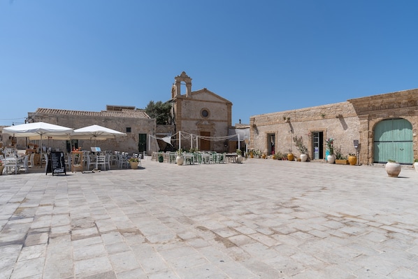 A courtyard with stone flooring, outdoor seating, and a building with a bell tower.