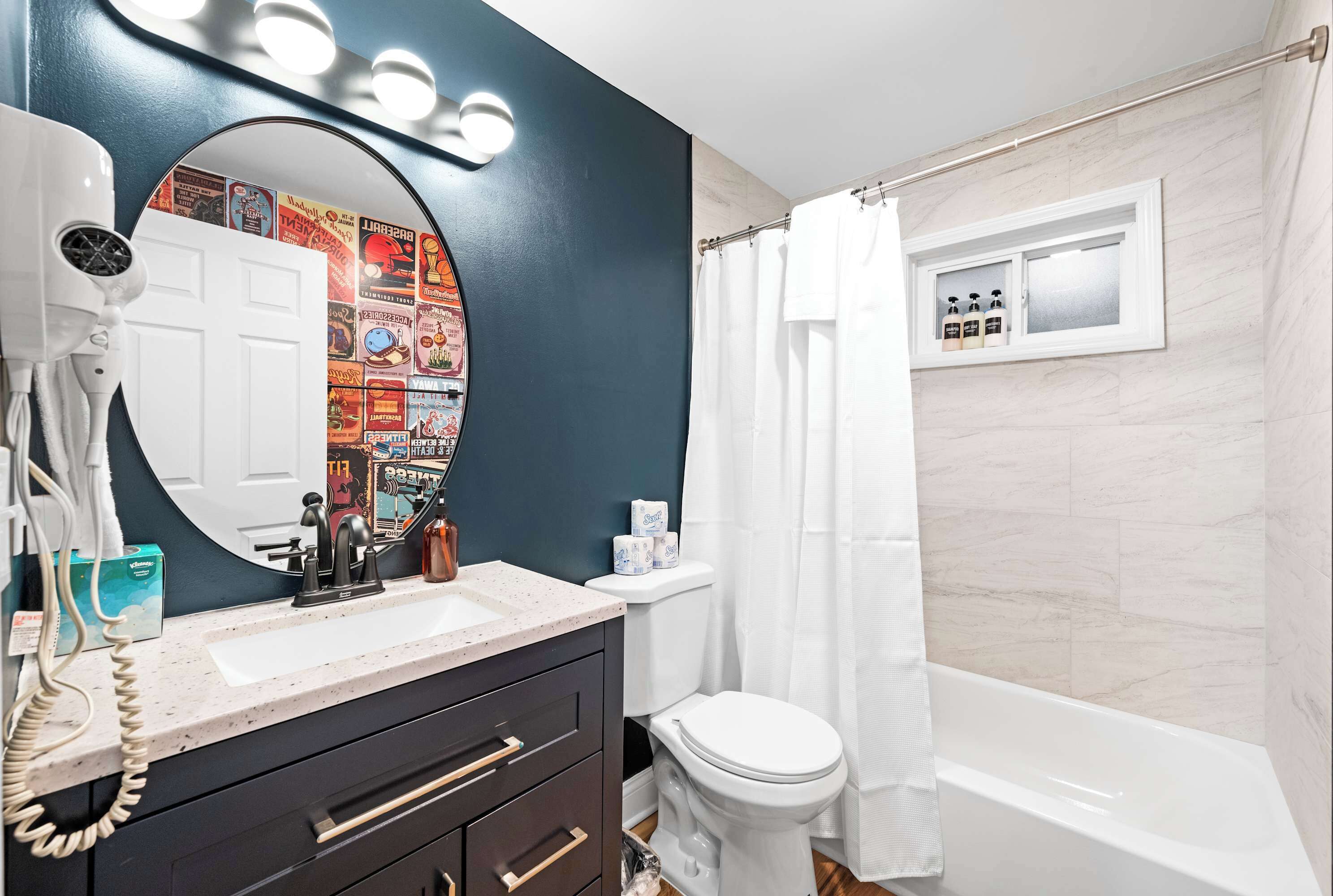 Une salle de bain avec un lavabo blanc, un meuble-lavabo foncé, un miroir rond avec éclairage intégré, des toilettes et une baignoire.