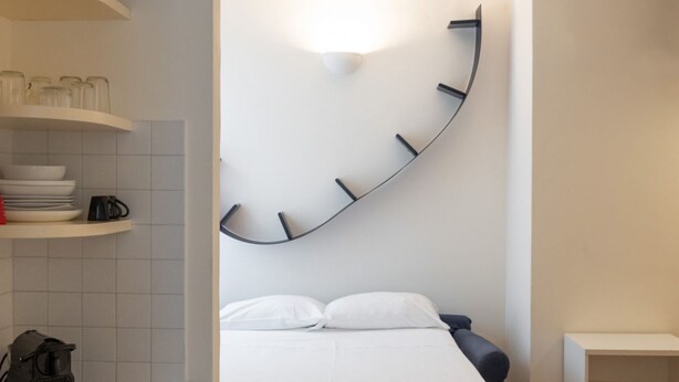 A modern hotel room with a curved wall clock, a bed with white linens, a small seating area, and a kitchenette with a coffee maker. - Studio apartment a stone's throw from CityLife (Milano)